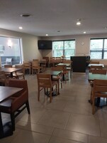 Un comedor con varias mesas y sillas, un televisor de pantalla plana montado en la pared y ventanas grandes con persianas.