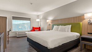 Ein modernes Hotelzimmer mit einem großen Bett, einem roten Sessel, einem Fenster mit Stadtblick und einer Wandlampe.