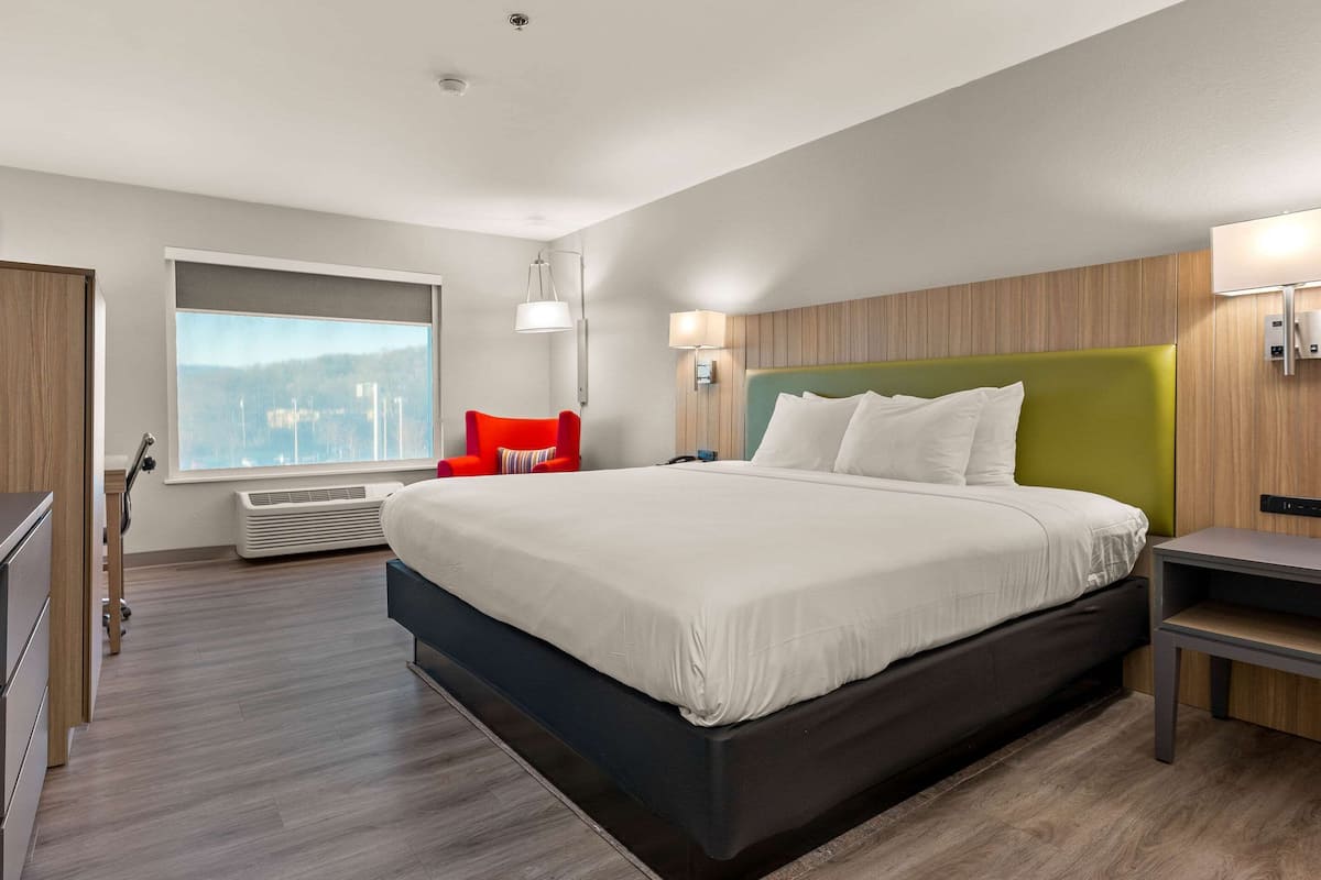 A modern hotel room with a large bed, a red chair, a window with a city view, and a wall-mounted lamp.