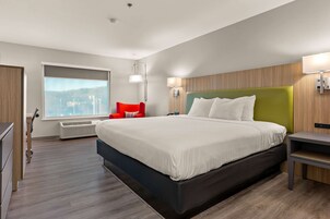 A modern hotel room with a large bed, a red chair, a window with a city view, and a wall-mounted lamp.