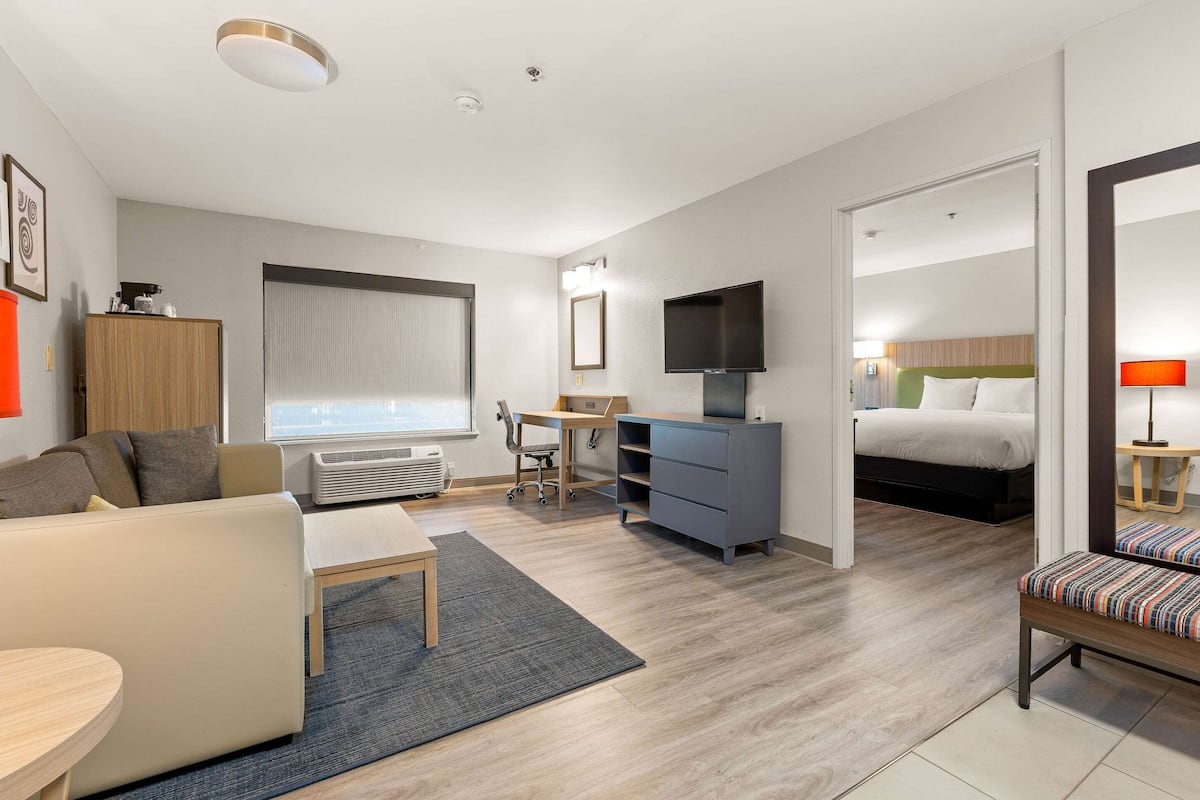 A modern hotel room with a sofa, a desk, a television, a bed, and a bathroom visible through an open door.