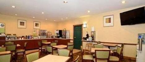 A hotel dining area with multiple tables and chairs, a counter with a cash register, and a flat-screen TV mounted on the wall.