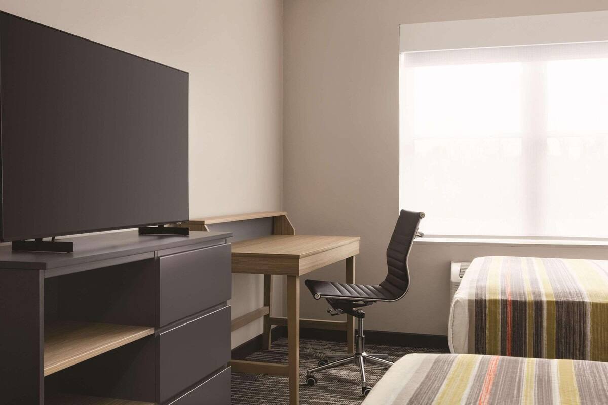 Une chambre d’hôtel équipée d’un bureau, d’une chaise, d’un lit avec une couvre-lit à rayures, d’un téléviseur posé sur un meuble et d’une armoire.