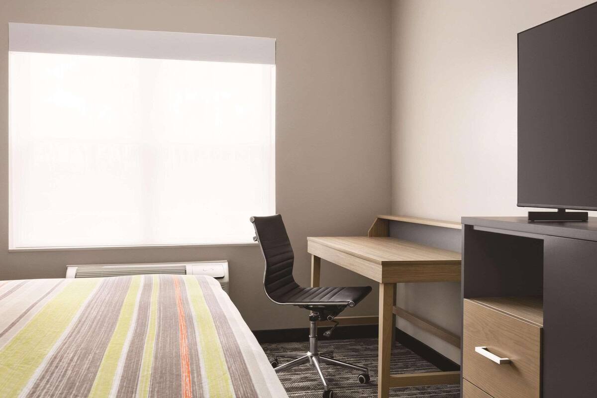 Une chambre d’hôtel avec un grand lit, un bureau avec une chaise, une télévision et une fenêtre avec des stores.