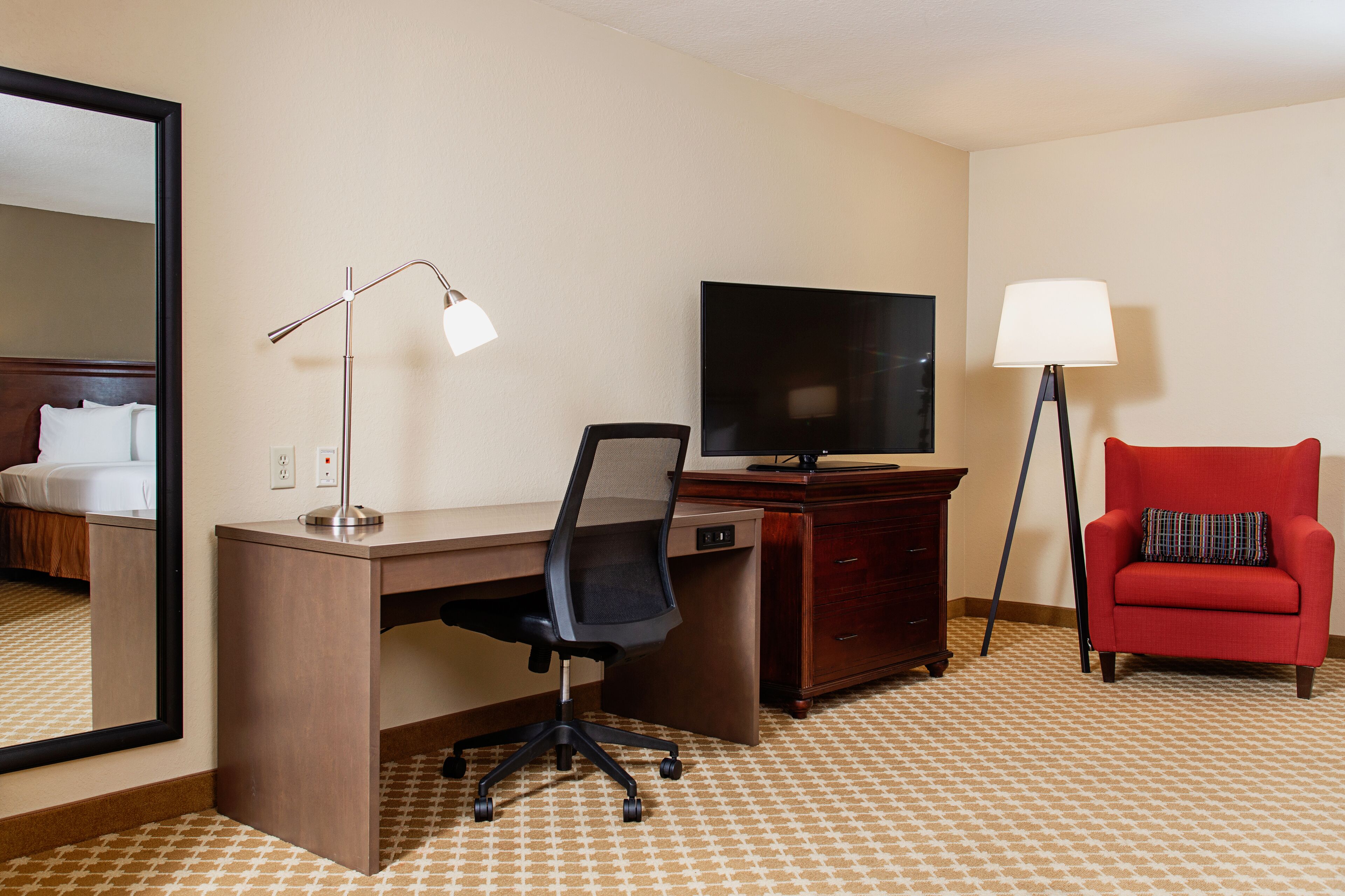 A hotel room with a desk, a television, a red armchair, and a large mirror.