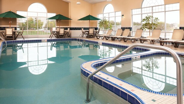 An indoor swimming pool with lounge chairs and umbrellas, surrounded by large windows. - Country Inn & Suites by Radisson, Milwaukee West (Brookfield), WI (Brookfield)