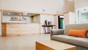 A modern reception area with a wooden reception desk, a grey sofa with an orange pillow, and a coffee machine. - Country Inn & Suites by Radisson, Milwaukee West (Brookfield), WI (Brookfield)