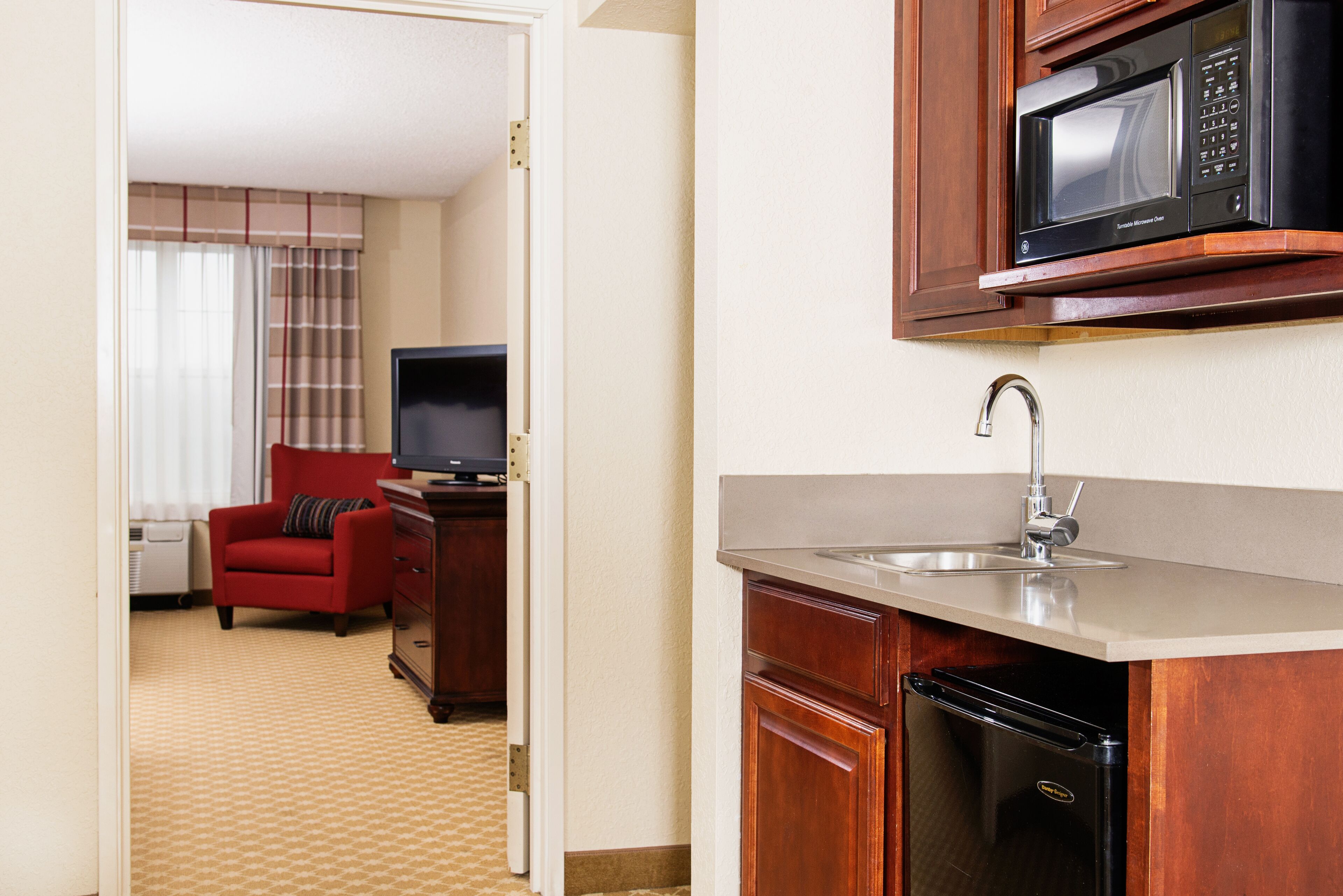 A compact hotel room with a kitchenette, a television, a red armchair, and a window with curtains.