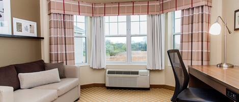 Una habitación con un sofá color crema, una silla de oficina negra, un escritorio y una ventana con cortinas escocesas.