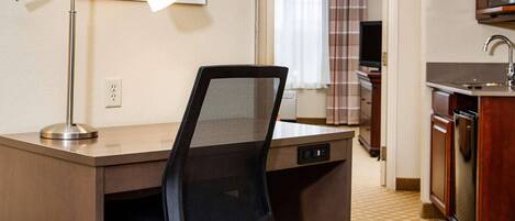 A compact hotel room with a desk, chair, kitchenette, and a framed picture on the wall.