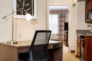 A compact hotel room with a desk, chair, kitchenette, and a framed picture on the wall.