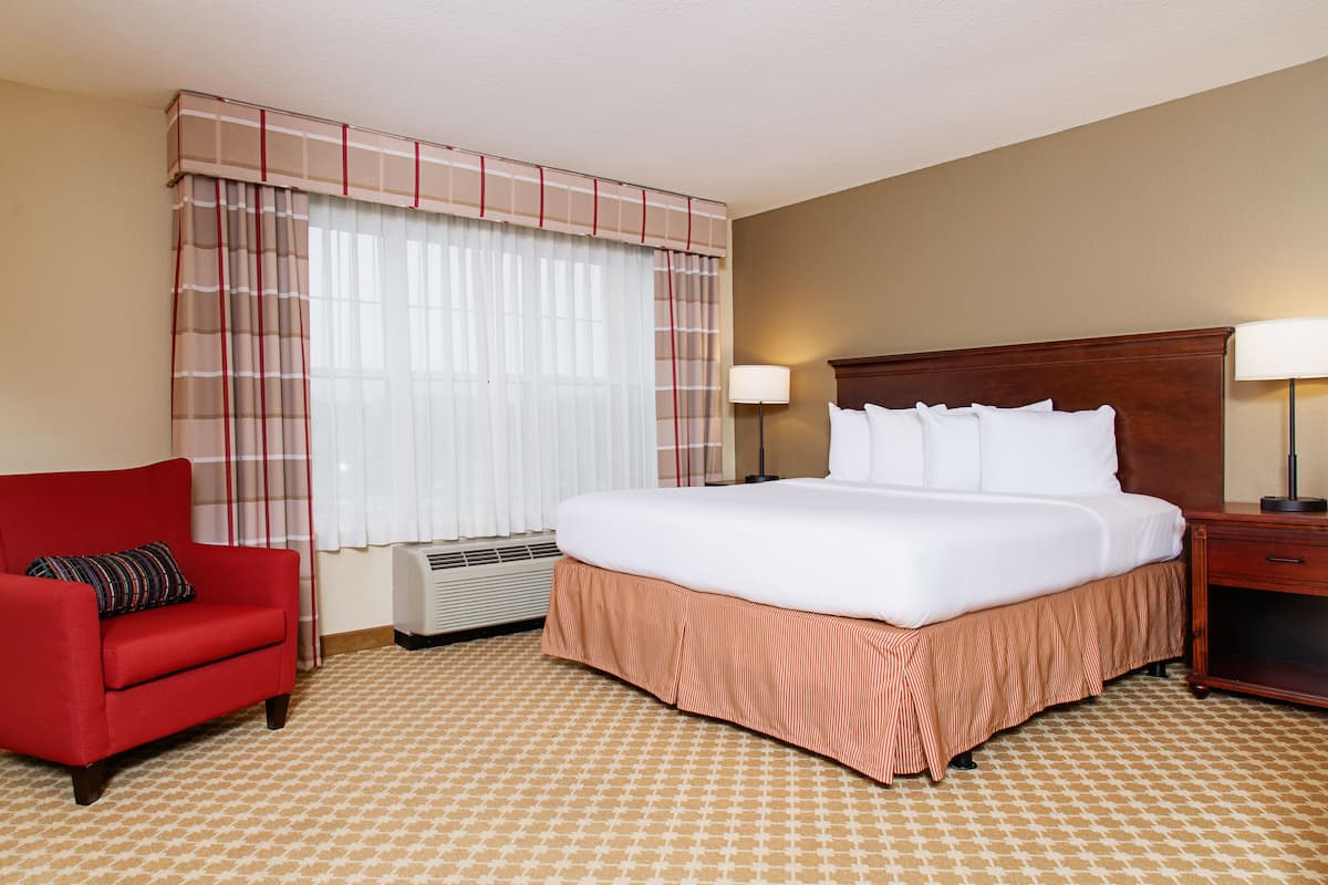 Une chambre d’hôtel avec un grand lit, un fauteuil rouge, une table de chevet avec une lampe et une fenêtre avec des rideaux.