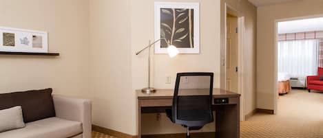 A hotel room with a sofa, desk, chair, and a framed leaf artwork on the wall.