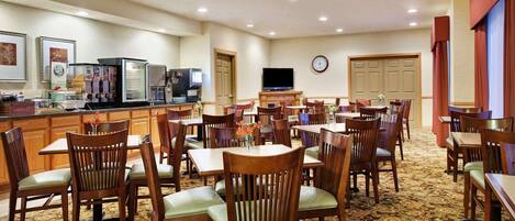 Un espace repas d’hôtel comprenant plusieurs tables et chaises, une machine à café et un téléviseur fixé au mur.