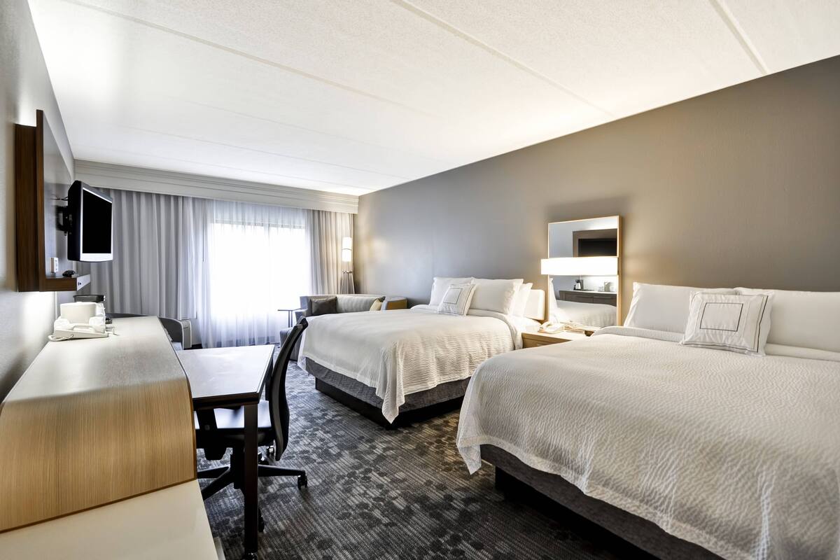 A hotel room with two beds, a desk, a TV, and a large window with curtains.