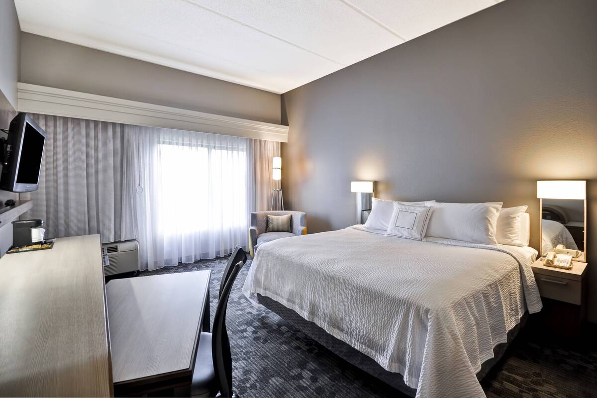 A hotel room with a large bed, a desk with a chair, a television, and a bedside table with a lamp.