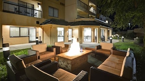 An outdoor seating area with wicker furniture and a central fire feature, surrounded by a building with balconies and large windows.