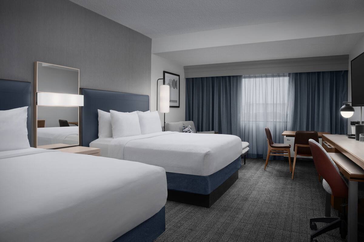 A hotel room with two beds, a desk, a chair, a lamp, and a large window with curtains.