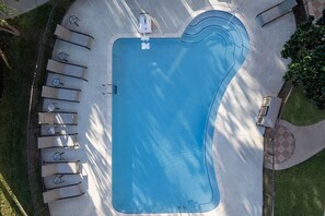 An aerial view of a swimming pool with a lounge area, surrounded by a paved deck and greenery.
