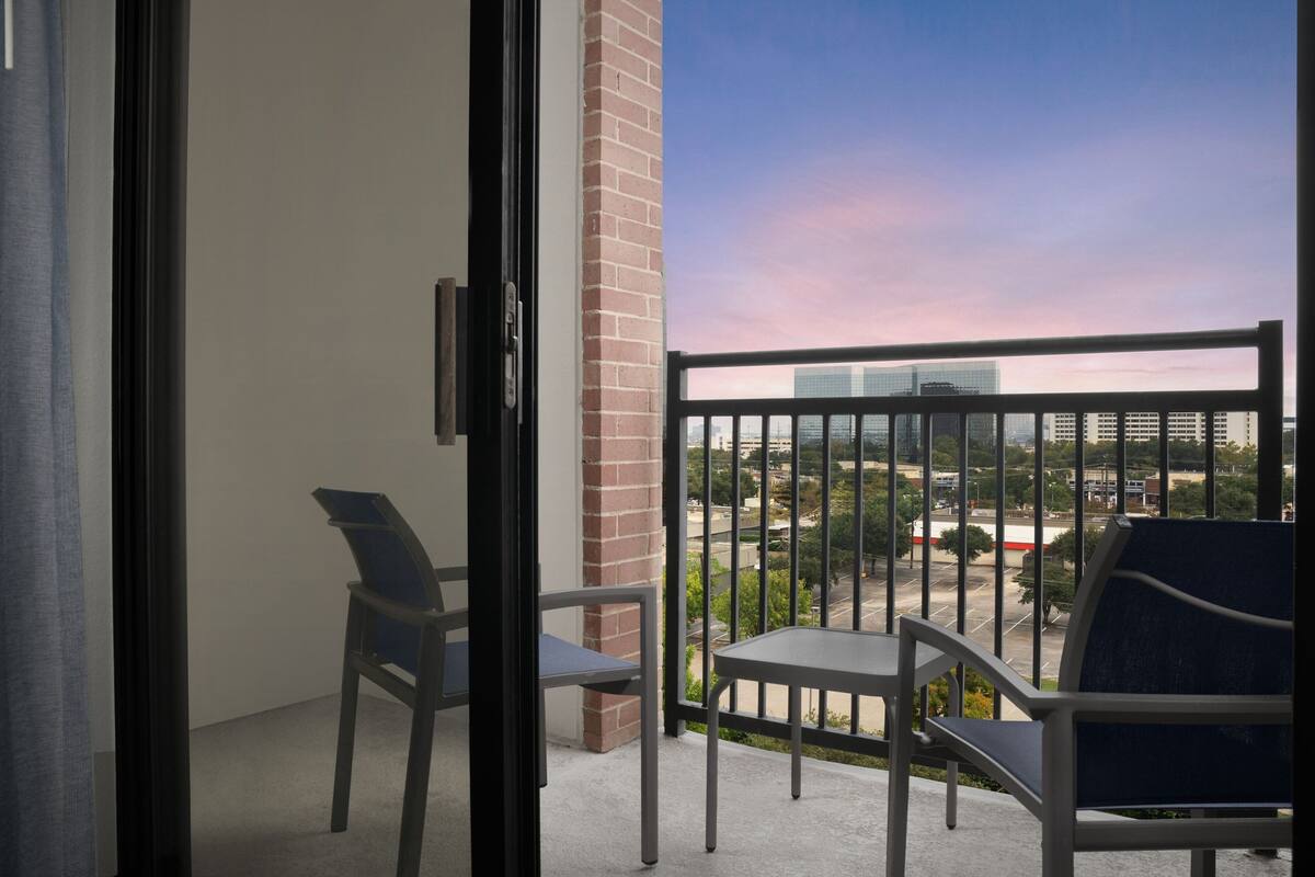 A balcony with two chairs and a table, overlooking a parking lot and buildings.