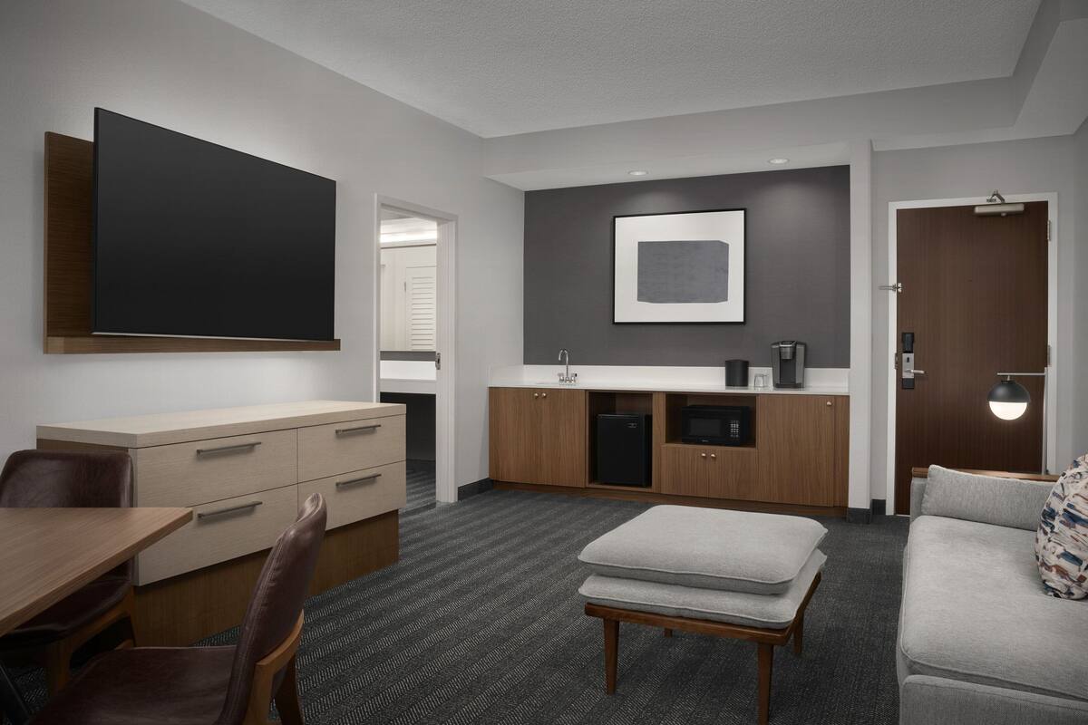 A modern hotel room with a flat-screen TV, a kitchenette, a sofa, and a dining area.