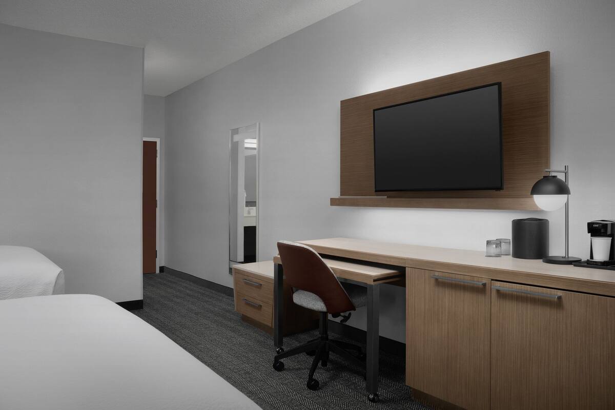 A hotel room with a bed, a desk with a chair, a flat-screen TV mounted on the wall, a lamp, and a coffee maker.