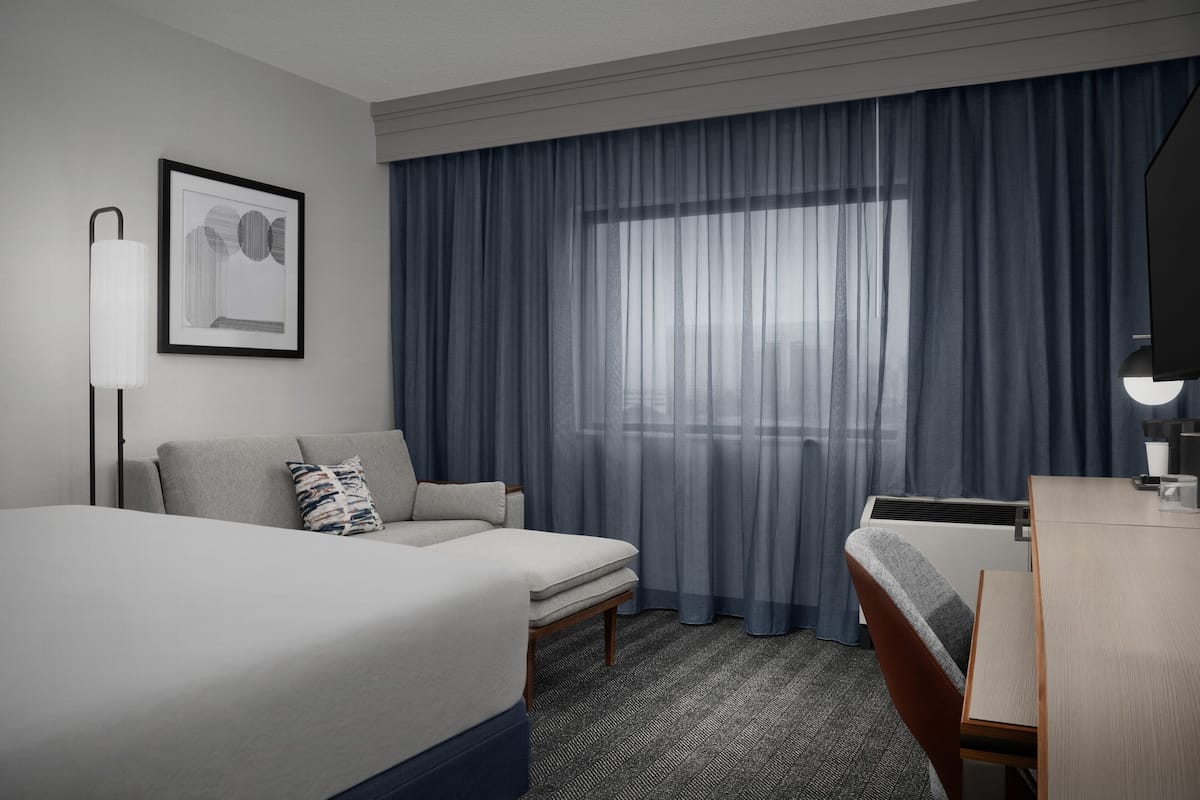 A hotel room with a bed, a sofa, a desk, a chair, a lamp, and a window with curtains.