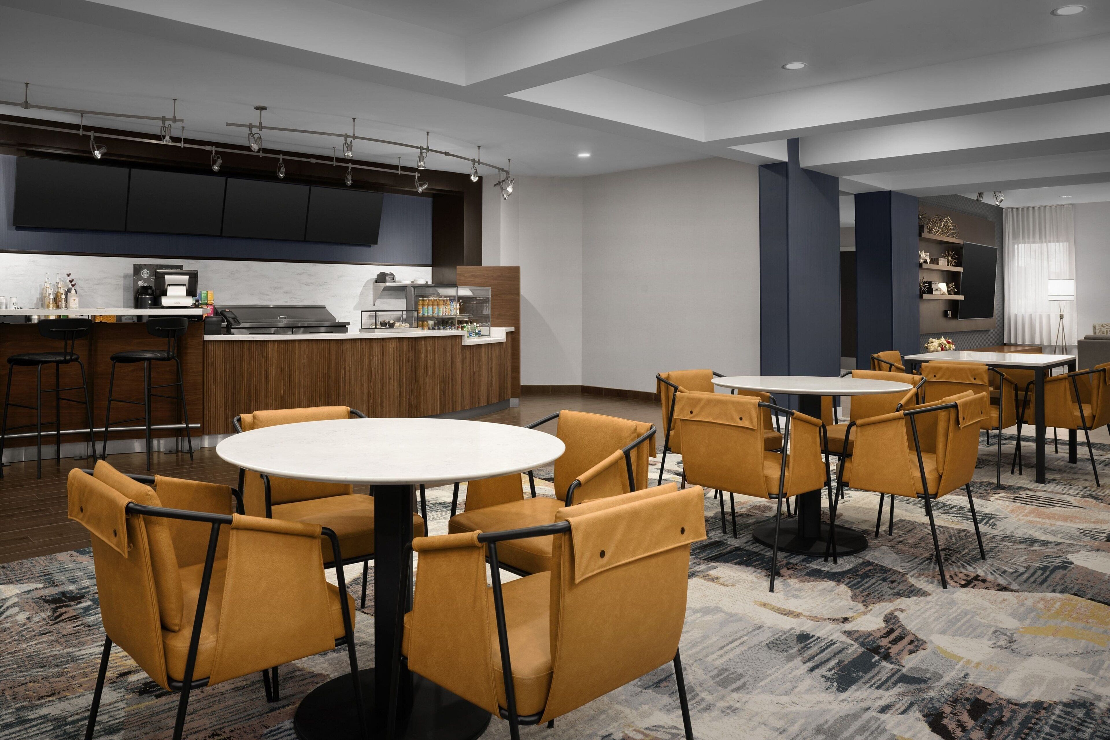 Un vestíbulo de hotel moderno con una mesa redonda central rodeada de sillas amarillas, una zona de bar con taburetes y una cocina al fondo.