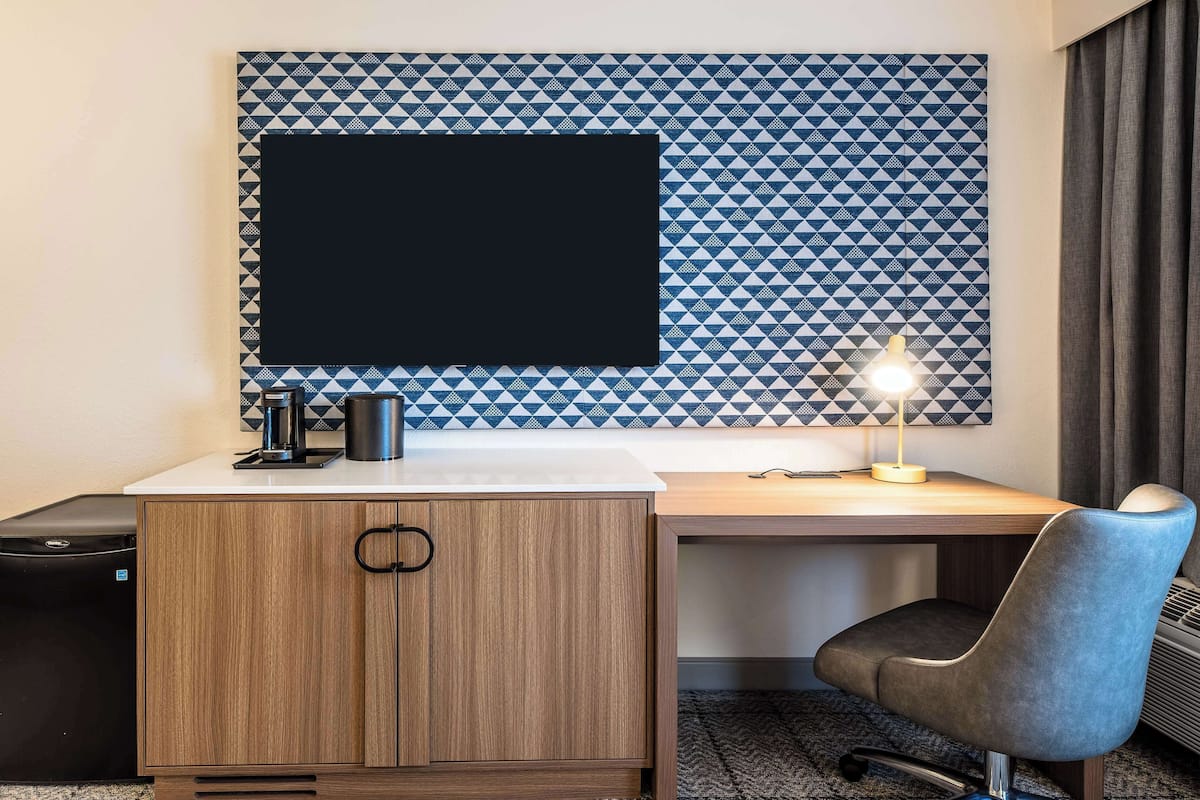 A hotel room with a wooden desk, a black monitor, a coffee maker, and a desk lamp.