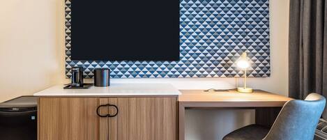 Une chambre d’hôtel avec un bureau en bois, un écran noir, une machine à café et une lampe de bureau.