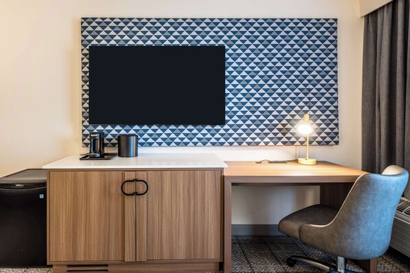 A hotel room with a wooden desk, a black monitor, a coffee maker, and a desk lamp.