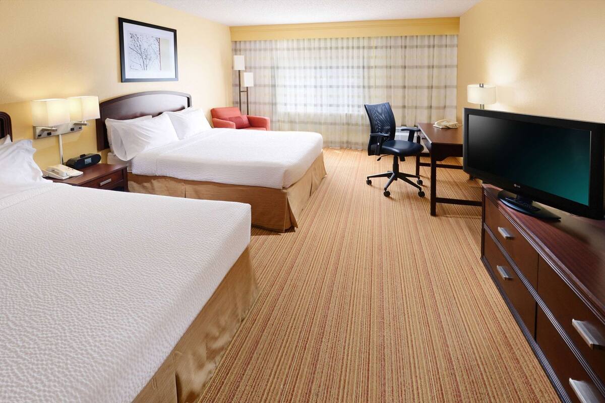 A hotel room with two beds, a desk with a TV, a chair, and a window with curtains.