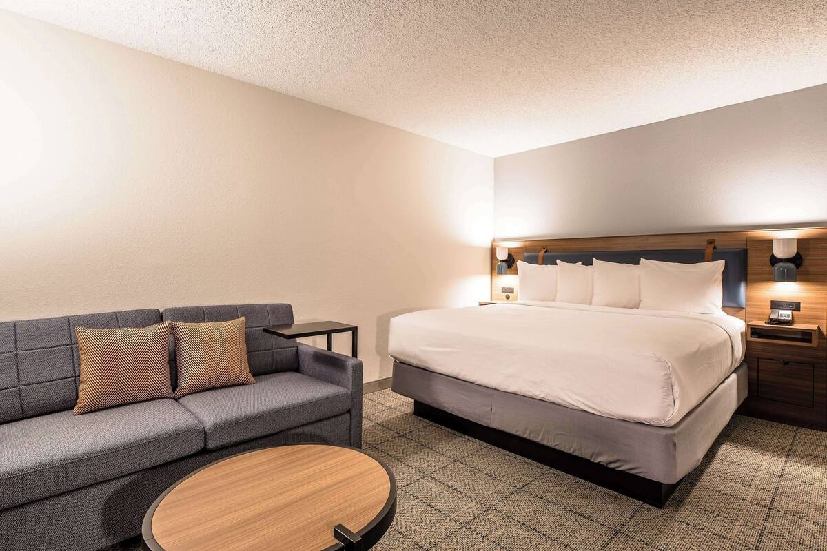 A hotel room with a bed, a sofa, a small table, and a nightstand with a lamp.
