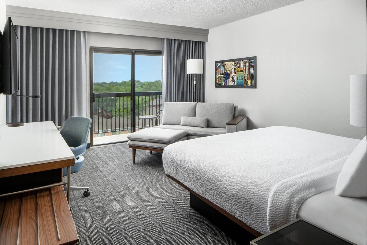 A hotel room with a bed, a desk, a chair, a sofa, a balcony with curtains, and a painting on the wall.