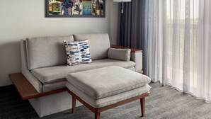 Una sala moderna con un sofá y reposapiés, una obra de arte enmarcada sobre el sofá, una lámpara y cortinas translúcidas.