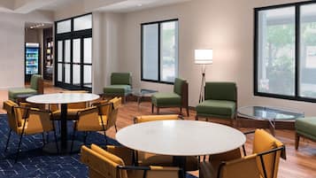 A modern hotel lobby with round tables and chairs, a vending machine, and large windows offering views of the outdoors.