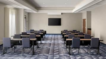 A conference room with a geometric patterned floor, rows of tables and chairs, a large black screen on the wall, and a door in the background.
