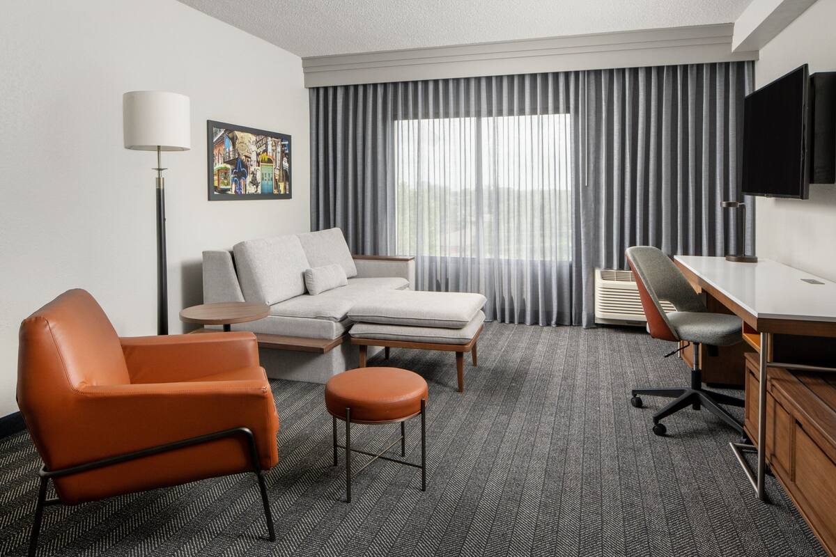 A hotel room with a sofa, armchair, ottoman, desk, chair, TV, and a painting on the wall.