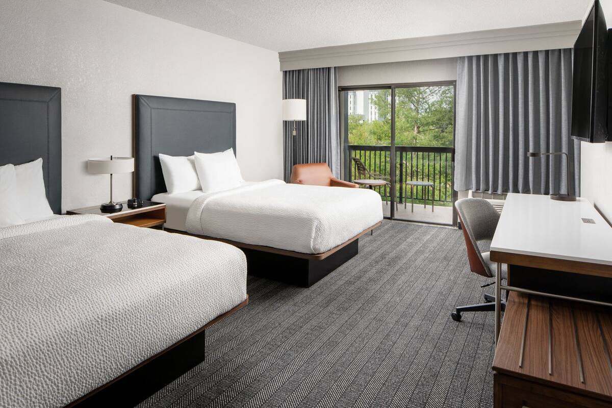 A hotel room with two beds, a desk, a chair, a TV, and a balcony with curtains.