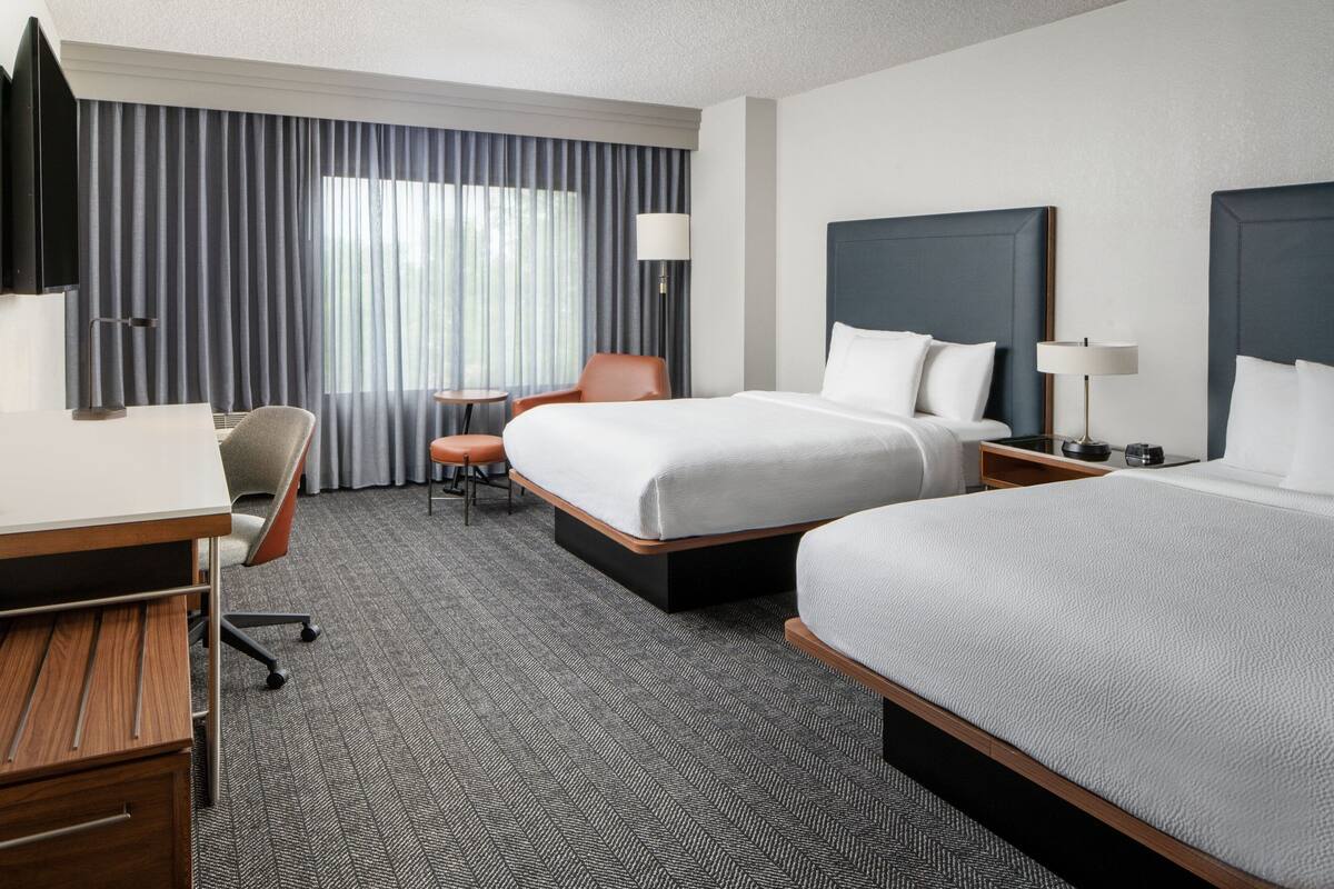 A hotel room with two beds, a desk, a chair, a TV, and a window with curtains.