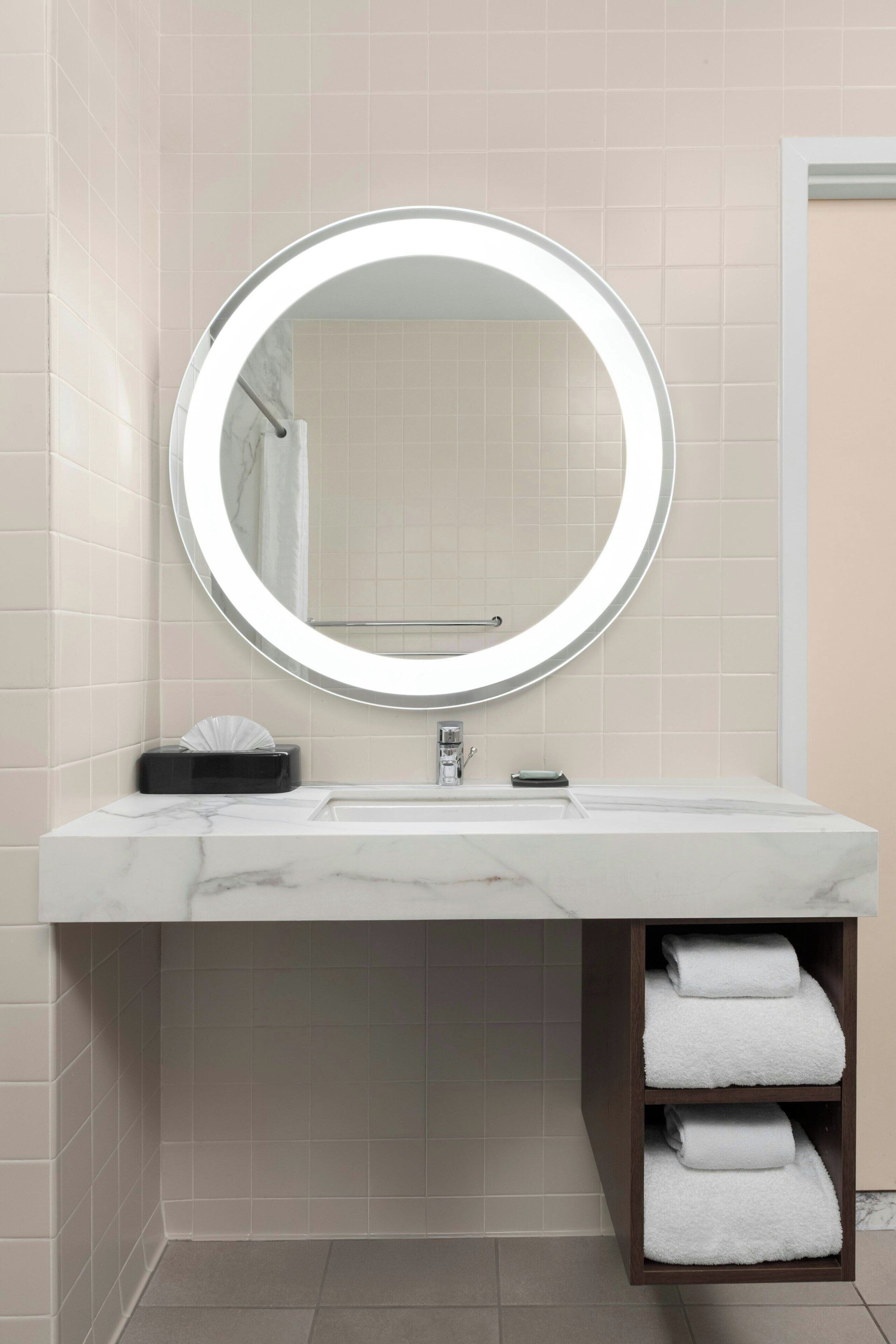 A bathroom with a round mirror, white marble sink, and a towel rack with folded towels.