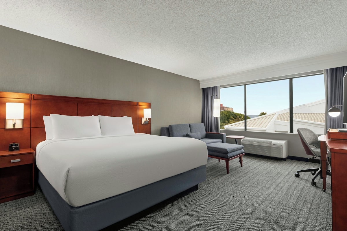 A hotel room with a large bed, a desk, a chair, and a view of the cityscape through large windows.