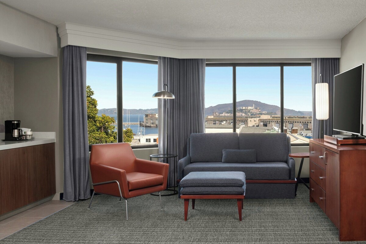 Une chambre d’hôtel équipée d’un canapé, d’un fauteuil, d’un pouf, d’une télévision et offrant une vue sur la ville et les montagnes.