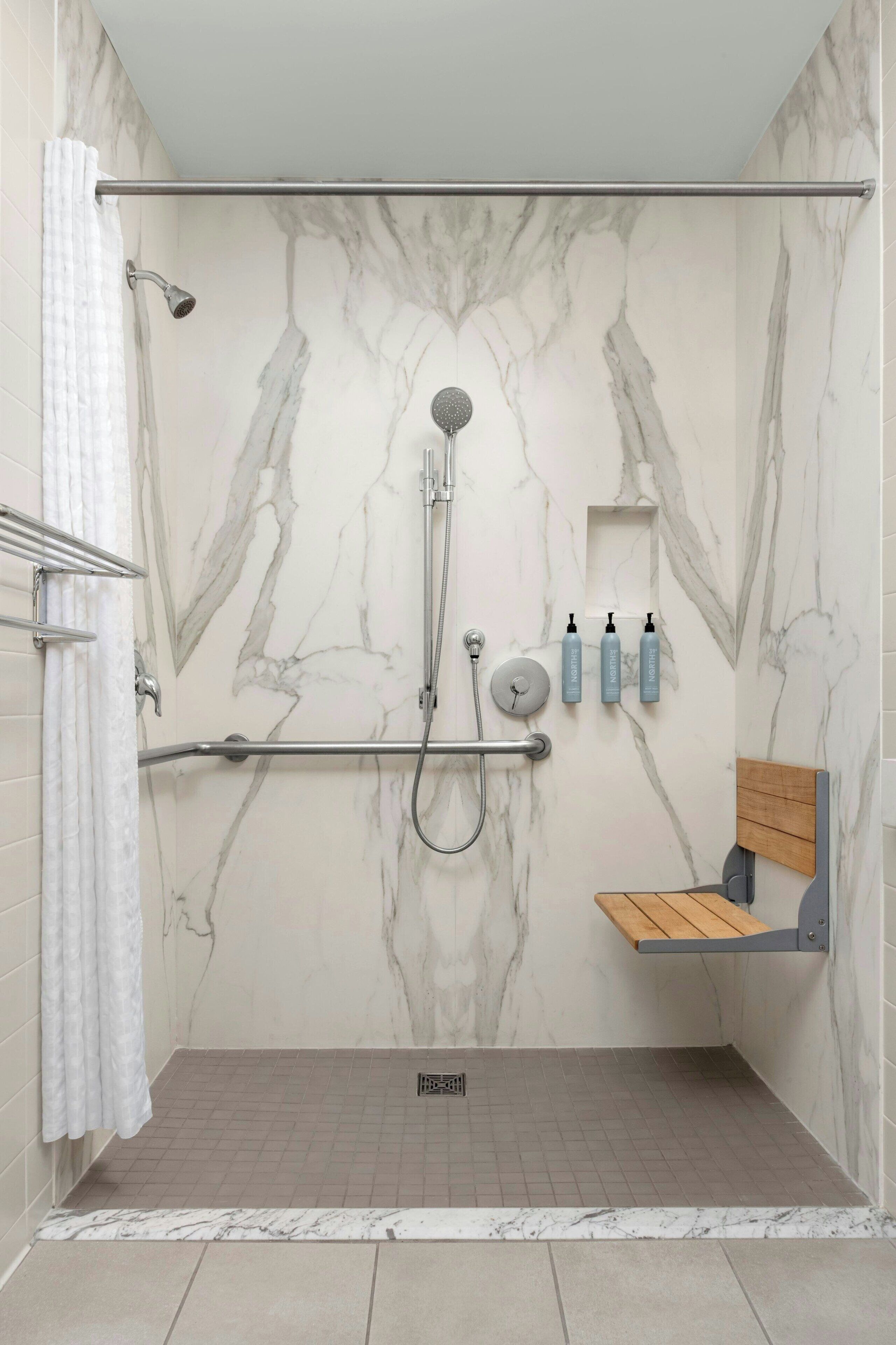 A modern shower with a marble wall, a grab bar, a handheld showerhead, and a shelf with toiletries.