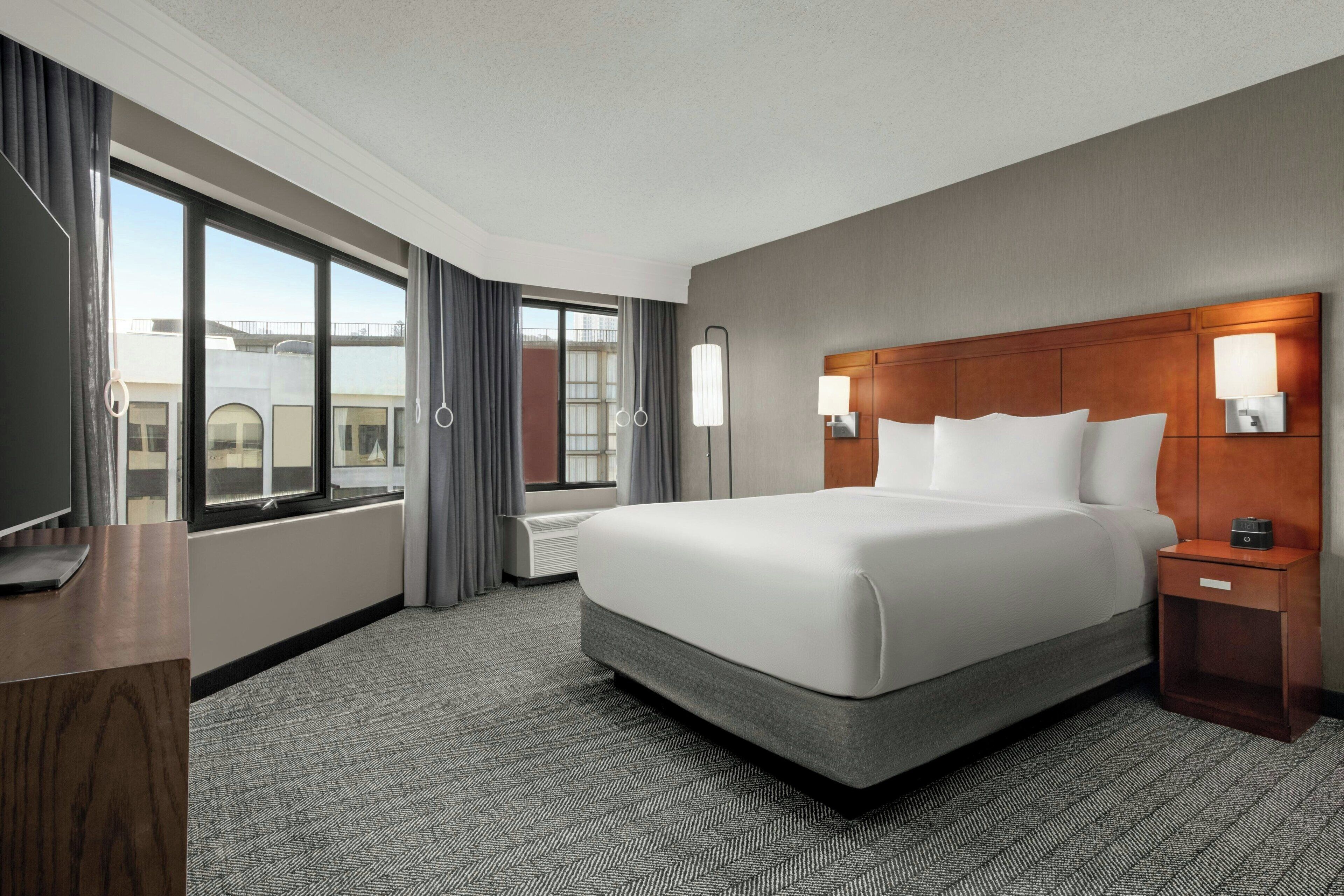A hotel room with a large bed, a desk, a chair, a nightstand, a lamp, and a view of the city through large windows.