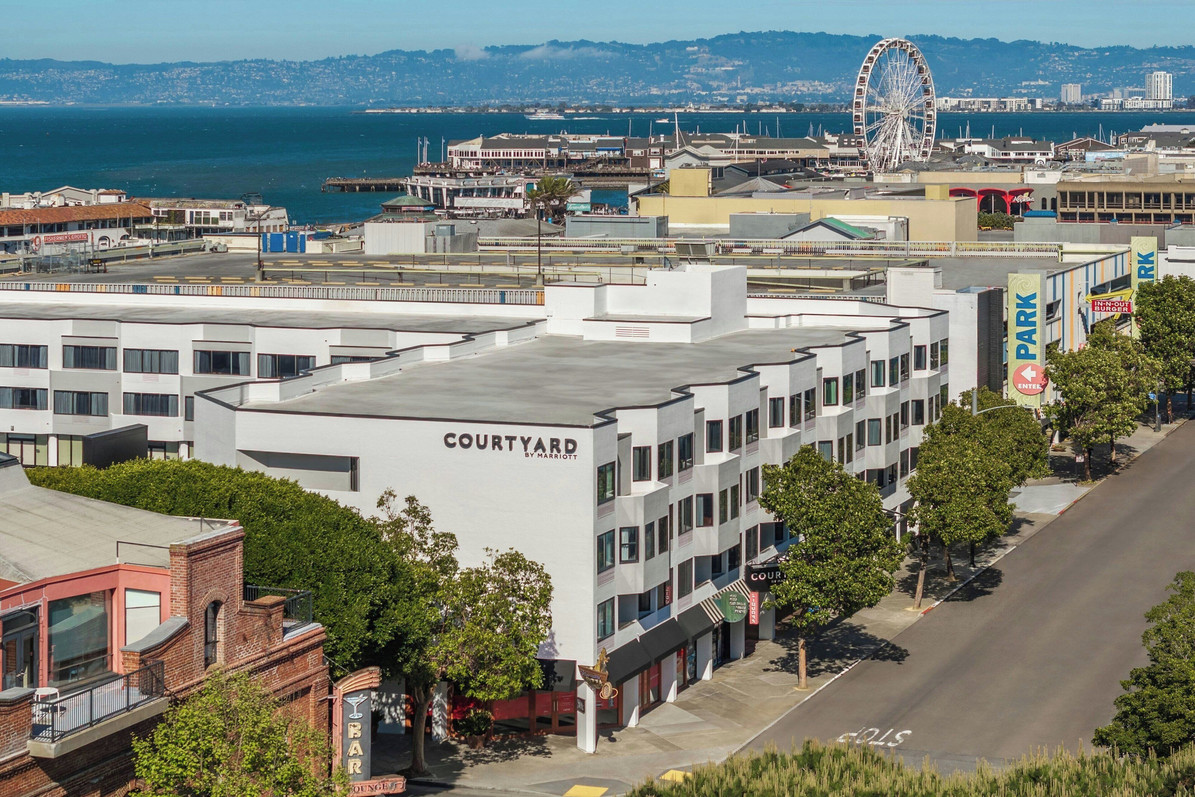 A Courtyard by Marriott hotel with a flat roof, located near a waterfront with a Ferris wheel in the background.