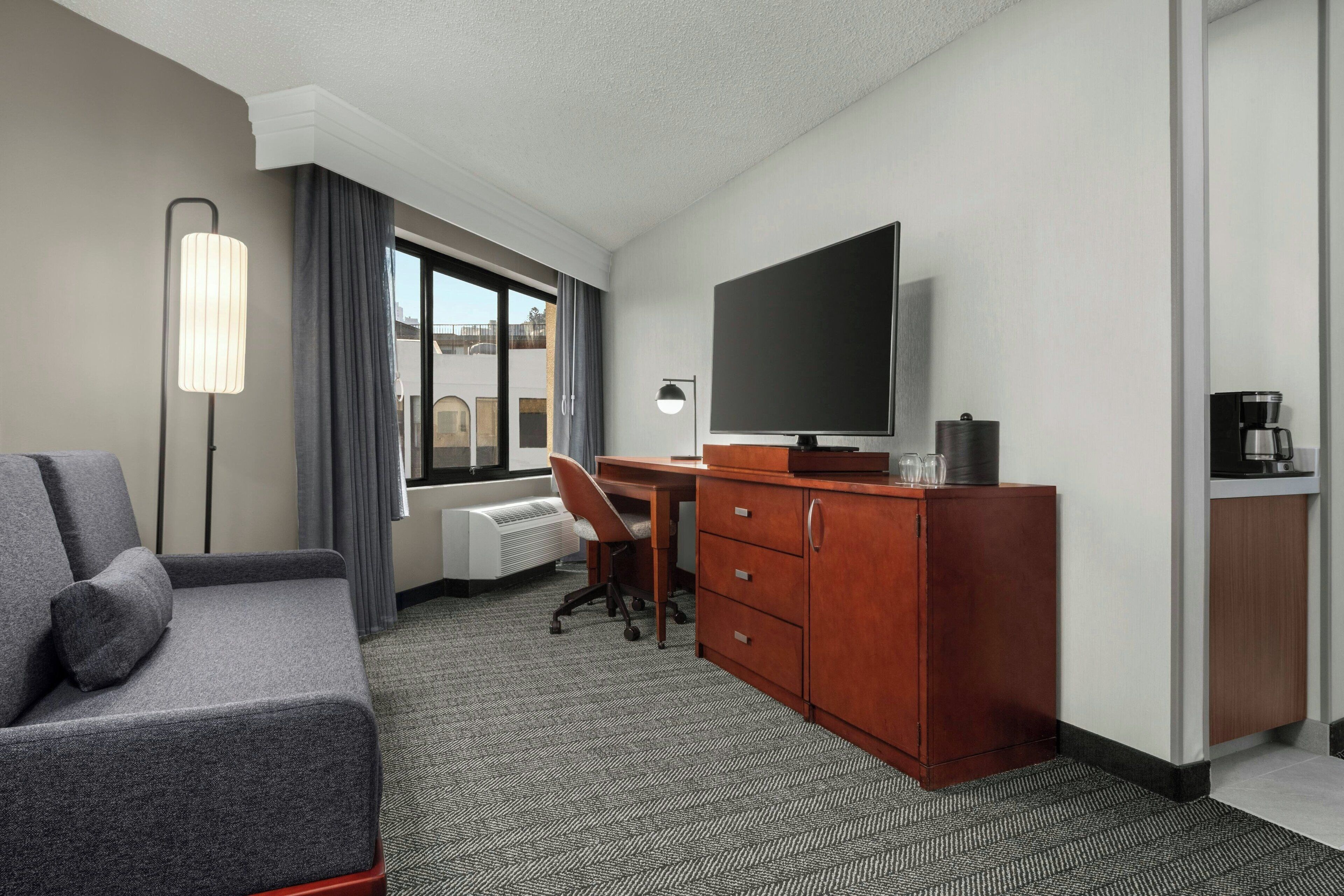 A hotel room with a gray sofa, a desk with a chair, a TV, a coffee maker, and a dresser.