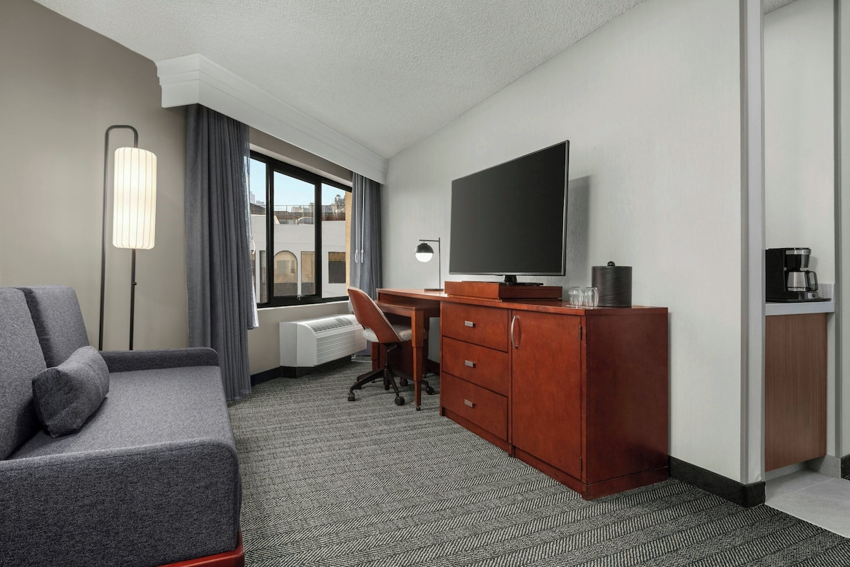 Une chambre d’hôtel avec un canapé gris, un bureau avec une chaise, une télévision, une machine à café et une commode.