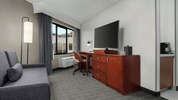 A hotel room with a gray sofa, a desk with a chair, a TV, a coffee maker, and a dresser.