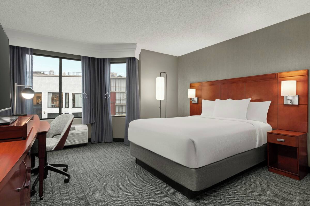 A hotel room with a large bed, a desk, a chair, and a view of the city through the window.